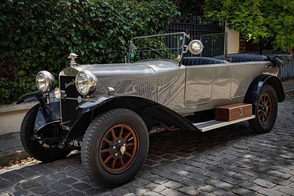 Voiture vintage argentée stationnée sur trottoir, devant végétation dense