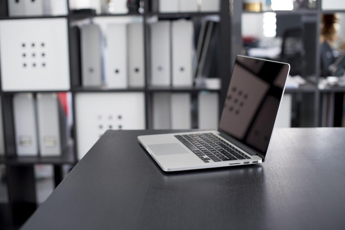 Un ordinateur portable ouvert sur un bureau noir, classeurs derrière