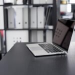 Un ordinateur portable ouvert sur un bureau noir, classeurs derrière