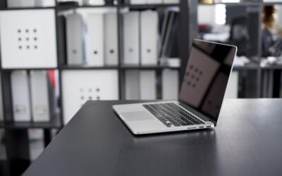 Un ordinateur portable ouvert sur un bureau noir, classeurs derrière