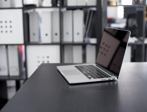 Un ordinateur portable ouvert sur un bureau noir, classeurs derrière
