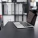 Un ordinateur portable ouvert sur un bureau noir, classeurs derrière